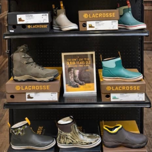 A retail shoe display features various LaCrosse boots and shoes on black shelves. Styles range from rugged to casual. A poster promotes durability.